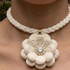 Peut inclure: Collier tressé de couleur crème avec un grand pendentif en forme de fleur. Le pendentif présente un diamant central entouré de petites pierres, serti dans un métal doré. Un bijou remarquable.