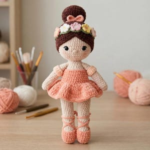 Puede incluir: Una muñeca de ganchillo con cabello castaño oscuro recogido en un moño, adornado con una diadema floral con flores rosas, blancas y amarillas. La muñeca viste un vestido color melocotón con falda de volantes y zapatillas de ballet a juego.
