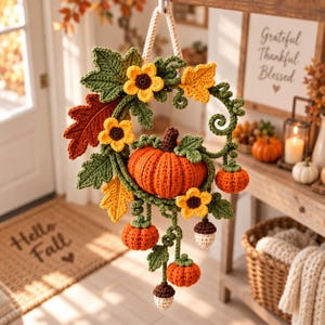Könnte beinhalten: Ein gehäkelter Wandbehang mit einem zentralen orangefarbenen Kürbis, kleineren Kürbissen, braunen Eicheln, gelben Blumen und grünen Blättern. Das Stück ist an einem dicken cremefarbenen Seil befestigt und weist detaillierte Garnstrukturen auf.