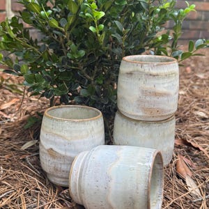 May include: Four cylindrical ceramic cups with a speckled off-white and tan glaze. The cups feature a smooth, rounded body with a slightly flared rim and a subtle horizontal ridge near the base, showcasing a natural, earthy color variation.