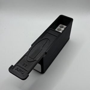 May include: Black rectangular container with a hinged lid, featuring the text "22 ARC." The lid has a raised bullet design. Inside, there are white cylindrical holders. The container is designed to hold small objects.