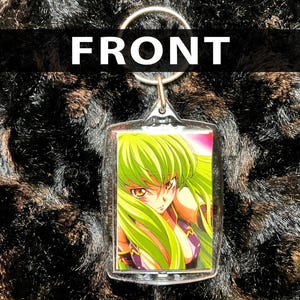 Może przedstawiać: Przezroczysty akrylowy brelok do kluczy z kolorową postacią z anime z zielonymi włosami i fioletowym strojem. Słowo "FRONT" jest wydrukowane na górze na biało. Brelok jest przymocowany do srebrnego metalowego pierścienia.