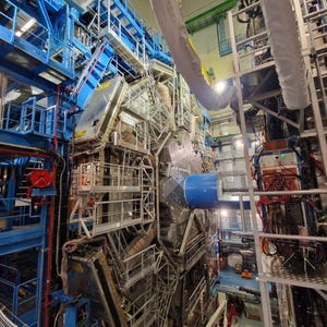 May include: An interior shot of a large scientific instrument, possibly a particle detector. The structure is a mix of blue, silver, and grey components, with many pipes, cables, and platforms. The scene is well-lit, suggesting a laboratory or research facility.