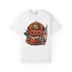 May include: White t-shirt featuring a detailed graphic of the CCCP emblem, including a red star, hammer and sickle, wheat, and a rocket. The design incorporates a tank, soldiers, and the word "CCCP" in red. The shirt has a crew neck.