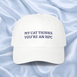 Puede incluir: Gorra de béisbol blanca con visera curva. La gorra presenta el texto "MY CAT THINKS YOU'RE AN NPC" en letras azul oscuro. El fondo es azul claro y sedoso.