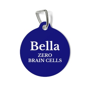 May include: A navy blue, circular pet tag with a silver-colored metal loop. The tag features the name "Bella" in white, above the text "ZERO BRAIN CELLS" in white, centered on the tag.