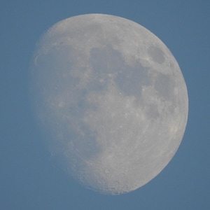 May include: A close-up of the moon in the sky. The moon is a pale gray color with visible craters and textures. The background is a clear, light blue sky.