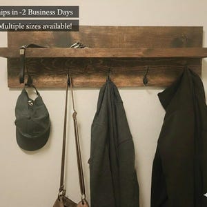 May include: A rustic, dark-stained wooden wall shelf with a top shelf and a lower shelf with three metal hooks. A green cap, a brown purse, and two dark-colored jackets hang from the hooks. The text "Multiple sizes available!" is visible.