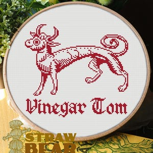 May include: A circular wooden embroidery hoop containing white fabric with a red cross-stitch design of a mythical creature with horns and a curled tail. Below the creature, the text Vinegar Tom is stitched in a red gothic-style font.