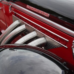 Puede incluir: Primer plano de un coche clásico rojo y negro. El coche presenta detalles cromados, incluyendo los faros, los tubos laterales y los adornos. El capó es negro y la carrocería es de un rojo vibrante.