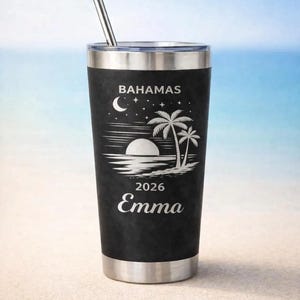 Peut inclure: Gobelet de voyage noir avec un bord et une paille argentés. Le gobelet présente un graphisme blanc d'un coucher de soleil, de palmiers et le mot "BAHAMAS". En dessous, les chiffres "2026" et le nom "Emma".