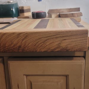 Op de afbeelding: Een houten snijplank met een lichtbruin oppervlak en een donkerbruine streep. De plank is gemaakt van meerdere stukken hout en ligt op een lichtbruine kast. Andere stukken hout en gereedschap staan op de achtergrond.