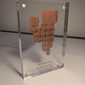 以下が含まれることがあります： 古代のパピルス、P52、ヨハネによる福音書18章31-33節と37-38節、125-150 ADの断片が透明なアクリルケースに展示されています。 パピルスは薄茶色で、黒い手書きのテキストが書かれています。 ケースは小さな透明な留め具で固定されています。