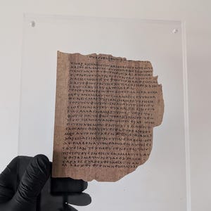May include: A fragment of ancient papyrus featuring handwritten Greek text in dark ink, preserved between two clear acrylic sheets. The papyrus has a rough, uneven edge on the right side and a light brown, fibrous texture throughout the surface.