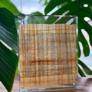 May include: A rectangular piece of light brown papyrus featuring dense columns of black Greek text, encased within a clear acrylic frame held together by four silver-toned metal corner screws, displaying a fibrous texture and uneven edges.