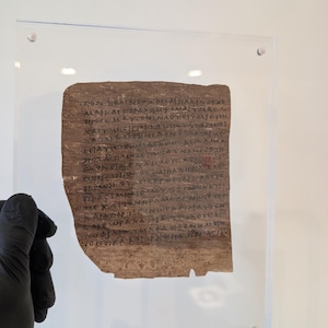 Può includere: Un frammento rettangolare di papiro antico con scrittura a mano in inchiostro marrone scuro in caratteri greci. Il papiro è montato tra due lastre di acrilico trasparente tenute insieme da quattro viti argentate agli angoli.