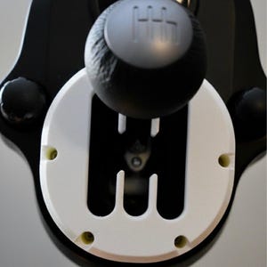 以下が含まれることがあります： ゲームコントローラーの黒と白のギアシフトのクローズアップ。 ギアシフトノブは、テクスチャ加工された表面とシルバーのギアパターンを備えた黒色です。 白いベースにはギアシフトパターンがあります。