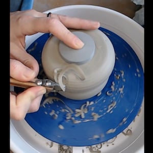 May include: A potter uses a trimming tool to shape a ceramic piece on a pottery wheel. The piece is light gray, with a lid and a handle being formed. The wheel is blue, and the hands are visible as they work on the pottery.