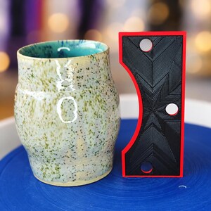 May include: A speckled ceramic mug with a light cream and green exterior and a turquoise interior, positioned next to a rectangular black tool with a red border, featuring a curved side and three circular holes in a geometric pattern.