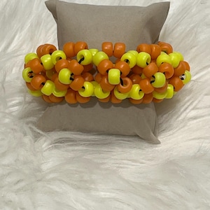 May include: A beaded bracelet featuring a mix of orange and yellow beads. The bracelet is made of small, round beads strung together, creating a chunky, textured look. The bracelet is displayed on a tan pillow against a white, fluffy background.