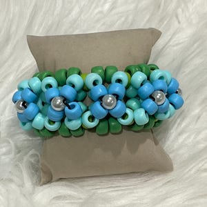 May include: A beaded bracelet featuring a floral design. The bracelet is composed of blue, light blue, and green beads, with silver-colored beads at the center of each flower. The bracelet is resting on a beige pillow.