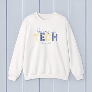 May include: A white crewneck sweatshirt with the text "Georgia TECH" in navy and yellow. The word "TECH" features a yellow and blue gingham pattern. The phrase "Yellow Jackets" is below the main text.