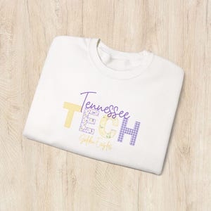 May include: White t-shirt with the words "Tennessee TECH Golden Eagles" in yellow, purple, and light green. The letters have floral and checkered patterns. The shirt is folded on a light wood surface.