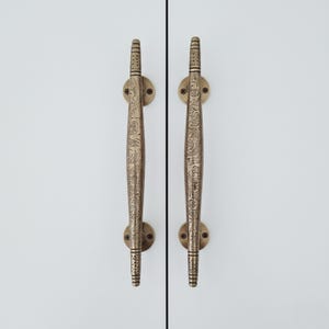 Könnte beinhalten: Zwei antike Messing-Schrankgriffe mit aufwendigen floralen Mustern. Jeder Griff ist ein langer, gebogener Stab mit dekorativen Details und einer kreisförmigen Basis zur Montage. Die Griffe sind auf einer weißen Oberfläche montiert.