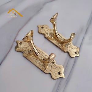 May include: Two ornate, gold-colored wall hooks. Each hook features a decorative, rectangular backplate with floral detailing and a curved hook. The hooks are made of metal and are designed for hanging items. The logo "House Craft MA" is in the top left corner.