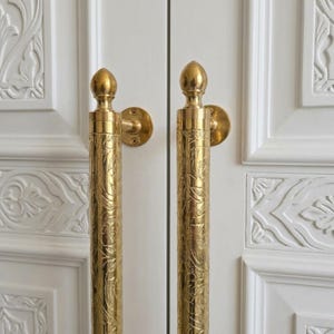 May include: Two vertical brass door handles with a polished finish and intricate engraved floral patterns. Each handle features a rounded finial at the top and a circular mounting plate secured with screws, attached to white paneled double doors.