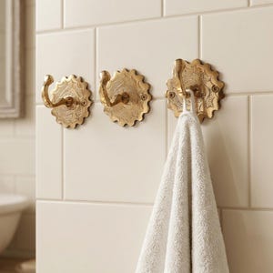 Op de afbeelding: Drie sierlijke, goudkleurige wandhaken met een bloemmotief zijn gemonteerd op witte vierkante tegels. Eén haak houdt een witte handdoek vast. De haken hebben een decoratieve, geschulpte rand, voor de badkamer.