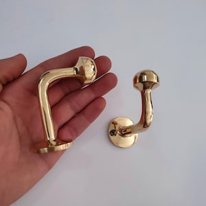 May include: Two polished brass coat hooks. One hook is held in a hand, the other is on a white surface. The hooks have a curved design with a rounded top and a circular base. The brass has a shiny, reflective finish.