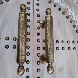 May include: Close-up of two ornate brass door handles with intricate floral patterns. The handles are mounted on a light-coloured wooden door, adorned with decorative studs. The door's design features carved details.
