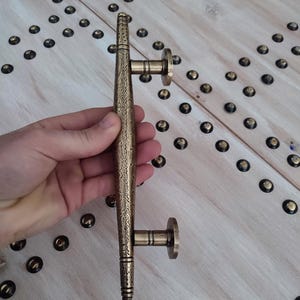 Può includere: Una maniglia per porta color bronzo con un design ornato e strutturato. La maniglia è tenuta da una mano, mostrando la sua dettagliata lavorazione. La maniglia ha due piastre di montaggio circolari. Lo sfondo è una superficie di legno chiaro con borchie decorative.