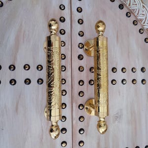 Könnte beinhalten: Zwei verzierte, goldfarbene Türgriffe mit aufwendigen floralen Designs. Die Griffe sind zylindrisch mit abgerundeter Ober- und Unterseite und an einer hellen Holztür befestigt. Die Tür ist mit kleinen, dunkel gefärbten Nieten verziert.