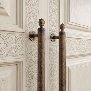 May include: Two vertical bronze-toned metal door handles with a cylindrical shape and a detailed engraved swirling pattern. Each handle features a rounded finial at the top and is mounted to a cream-colored door with matching carved scrollwork.