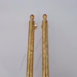 May include: Two ornate, gold-colored curtain rods with a detailed, engraved floral pattern. Each rod has a rounded finial at the top and a mounting bracket. The rods are displayed against a plain white background.