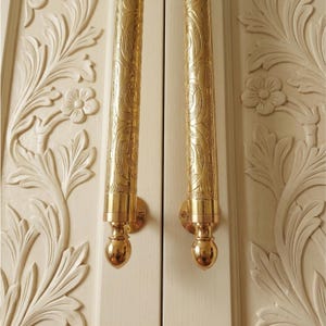 Puede incluir: Dos manijas de puerta verticales de metal dorado con intrincados patrones grabados y remates redondeados, montadas en un par de puertas de madera color blanco hueso con relieves tallados de flores y hojas en toda la superficie.