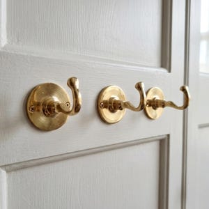 May include: Three brass wall hooks fixed to a light grey door. The hooks feature a traditional design with a circular base and a curved hook. The brass finish offers a warm, vintage aesthetic. Ideal for hanging coats, hats, or other items.
