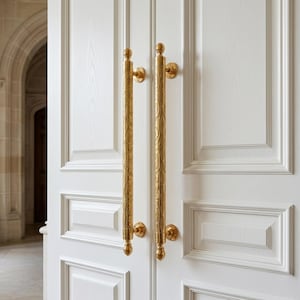 May include: A pair of white doors with ornate, gold-colored handles. The handles are long, cylindrical, and feature detailed floral patterns. The doors are paneled and set in a light-filled space.