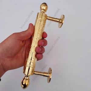 Pode incluir: Uma puxador de porta dourado com um design floral ornamentado. O puxador da porta é segurado em uma mão e apresenta uma alça cilíndrica com pináculos decorativos em cada extremidade. O texto "HouseCraftMA" é visível na imagem.