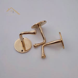 Puede incluir: Tres perchas de metal dorado. Dos ganchos tienen un brazo curvo y una base redonda, mientras que el tercero tiene un brazo recto y una base redonda. Los ganchos están diseñados para montaje en pared y probablemente se utilizan para colgar abrigos u otros artículos.