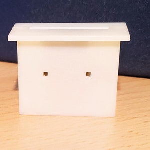 Puede incluir: Una pequeña caja rectangular blanquecina con una parte superior plana y dos agujeros cuadrados en la parte delantera. La caja está sobre una superficie de madera. El fondo es de color azul oscuro.