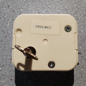May include: White plastic music box with a key wind-up mechanism. The box has a label that reads "FUNERAL MARCH".