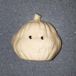 Puede incluir: Una calabaza de cerámica blanca con una cara tallada. La calabaza tiene dos pequeños agujeros para los ojos y una línea curva para la boca.