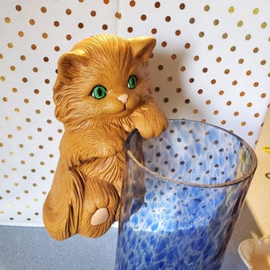 Op de afbeelding: Een keramiek beeldje van een bruine kat met groene ogen, geperst op de rand van een blauwe glazen vaas met een wit gevlekt patroon.