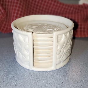 May include: A set of eight white ceramic coasters with a raised design of leaves and berries. The coasters are stacked in a holder with a similar design.