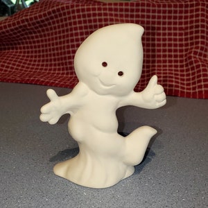 Puede incluir: Figurita de fantasma de cerámica blanca con una cara sonriente y pulgar hacia arriba. El fantasma tiene un cuerpo redondeado y una cola larga y fluida.