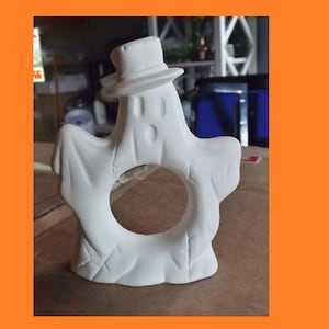 Könnte beinhalten: Ein weißer Keramik-Serviettenring in Form eines Gespenstes mit Hut und einem Loch in der Mitte für eine Serviette. Das Gespenst hat einen überraschten Ausdruck.