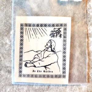 May include: A small, square, off-white image featuring a black and white illustration of a figure in a robe, looking upward. The image is framed by a decorative border and includes the text "In The Garden" at the bottom. The image is packaged in a clear plastic sleeve.